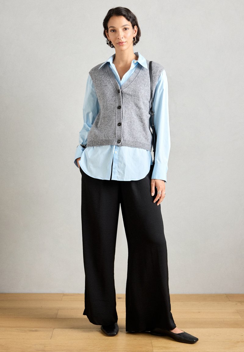 Grauer Strickweste über einem hellblauen Hemd mit Knöpfen, kombiniert mit schwarzen weiten Hosen und schwarzen Ballerinas. Glatte Texturen und klassischer Stil.