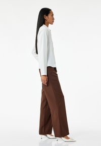 Camisa blanca de manga larga combinada con pantalones de pierna ancha marrones y tacones blancos puntiagudos. Tejido suave con un corte relajado y detalle de cuello.