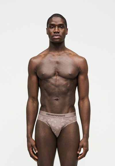 Μυώδης άνδρας με σκουρόχρωμο δέρμα στέκεται πρόσωπο με πρόσωπο, φορώντας μπεζ εσώρουχα Emporio Armani με ζώνη μέσης με μοτίβο λογότυπου σε λευκό φόντο.