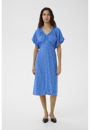 Mujer con un vestido midi azul con pequeños estampados florales, mangas cortas abullonadas y zapatos planos negros con punta, de pie contra un fondo liso.