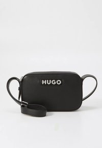 Musta nahkainen käsilaukku hopeisella "HUGO"-logolla, suorakulmainen muoto, vetoketjukiinnitys ja säädettävä olkahihna. Sileä pinta ja minimalistinen muotoilu.