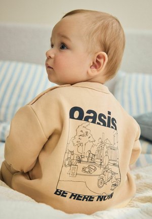 Dieťa oblečené v béžovej mikine s čiernou linkovou grafikou ulickej scény a textom "oasis" a "be here now" na zadnej strane.