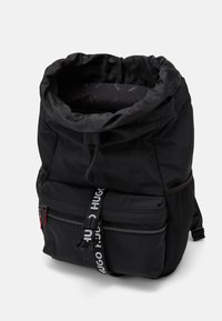 Zaino nero in nylon con apertura superiore a coulisse, tasca frontale con zip e tasca laterale in rete. Presenta una tracolla con logo "HUGO" e texture lisce.