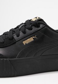 Detailní záběr na černou tenisku Puma s černými tkaničkami, zlatým logem Puma na boku a jazyku, a texturovanou podrážkou.
