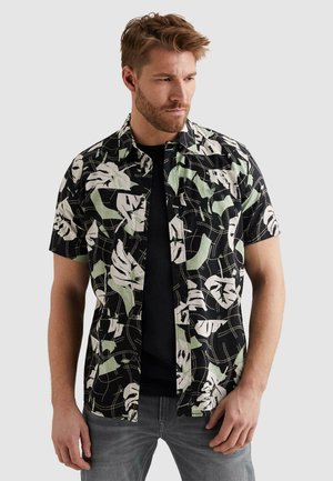 Mann trägt ein schwarzes Kurzarmhemd mit weißen und hellgrünen abstrakten Blattmustern, schwarzes Unterhemd und graue Jeans.