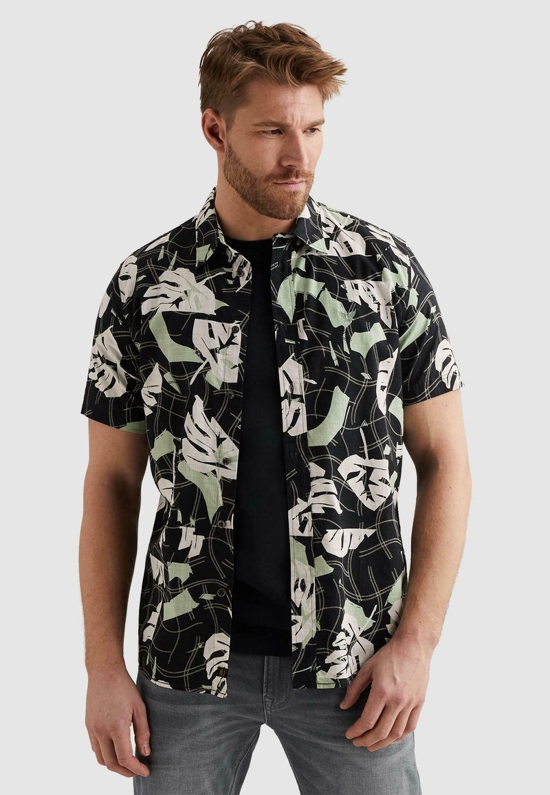 Homme portant une chemise noire à manches courtes avec des motifs abstraits de feuilles blanches et vert clair, un sous-t-shirt noir et un jean gris.