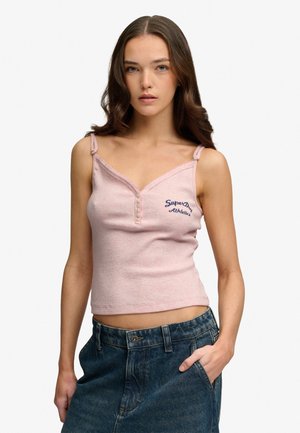 Jonge vrouw met golvend bruin haar die een roze tanktop met knopen draagt en loszittende blauwe spijkerbroek, met één hand in de zak.