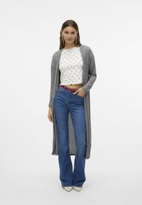 Ljusgrå lång cardigan över en vit crop top med blommönster. Utställda blåjeans och spetsiga vita klackar fulländar looken.