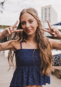 Marineblauw cropped top met gerimpelde textuur, dunne bandjes en een uitlopende kant zoom met oogjespatronen. Model maakt een vredesteken.