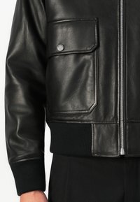 Manga de chaqueta de cuero negra y bolsillo delantero con tapa y cierre de presión metálico, cremallera frontal, puños y cinturilla acanalados, llevado sobre pantalones negros.
