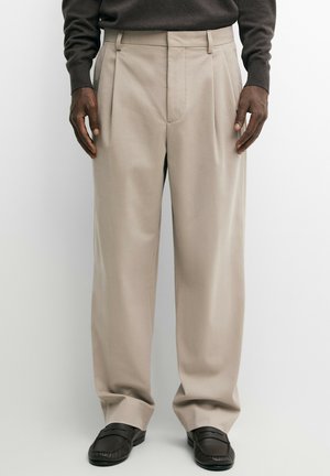 Mand iført beige plisserede bukser, mørkebrun sweater og sorte loafers stående mod en ensartet lys baggrund.
