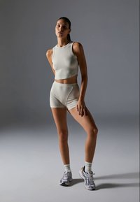 Junge Frau posiert vor grauem Hintergrund in einem hellgrauen ärmellosen Crop-Top, passenden hochgeschnittenen Shorts, weißen Socken und weißen Turnschuhen.