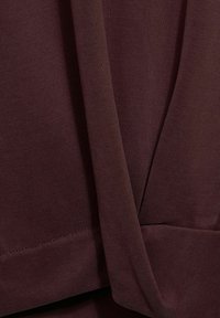 Bordowy materiałowy top z miękką, teksturowaną powierzchnią, o kroju drapowanym i wykończonych szwach. Materiał wydaje się elastyczny i wygodny.