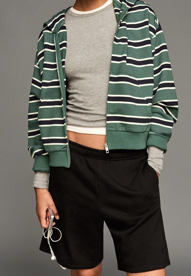 Personne portant un sweat à capuche zippé rayé vert et noir par-dessus un crop top gris et un short noir, tenant un téléphone avec des écouteurs blancs.