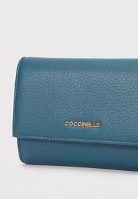 Cartera de piel texturizada azul con cierre de solapa y logotipo dorado "COCCINELLE" en la parte frontal.