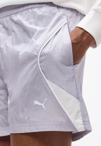 Χέρι στην τσέπη των αθλητικών σορτς Puma ανοιχτού γκρι χρώματος με λευκή λεπτομέρεια και λογότυπο στο αριστερό μηρό, φορεμένα με λευκό μπλουζάκι μακρυμάνικο.