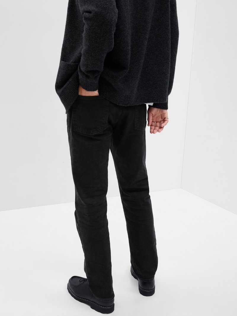 Pantalons en coton noir avec une coupe droite, poches arrière visibles et une finition texturée, associés à des chaussures noires.