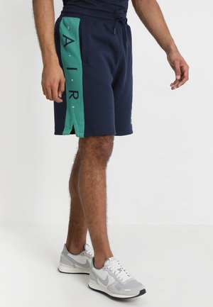 Homme portant un short bleu marine avec des panneaux latéraux turquoise étiquetés "NIKE AIR" et des baskets Nike grises, debout sur un fond blanc.