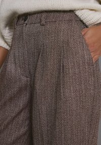 Pruun herringbone-mustriga püksid voldikute ja vöörihma aasadega, küljetaskudega ja tekstuurse kangaga.