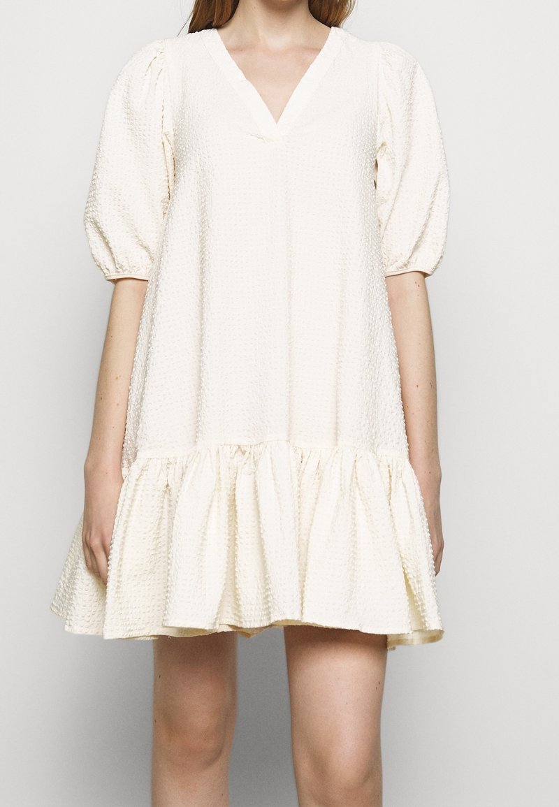 Frau in einem weißen, strukturierten Mini-Kleid mit Puffärmeln und einem gerafften Saum, vor einem schlichten, hellen Hintergrund stehend.