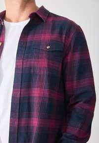 Camisa de franela en un plaid de azul oscuro y magenta, con cuello abotonado, dos bolsillos en el pecho y acentos de botones metálicos.