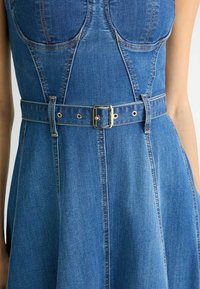 Jeanskleid in hellblauer Farbe mit tailliertem Oberteil, ausgestelltem Rock und breitem Gürtel. Verfügt über goldene Beschläge und doppelt genähte Nähte.