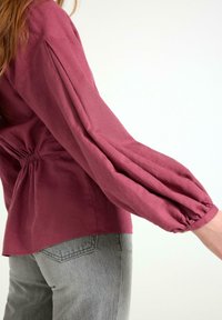 Langærmet top i burgunderrød med indsnævret talje og samlede ærmer. Komplementeret af en afslappet pasform og parret med lysegrå, tapered jeans.