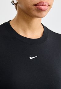 Svart bomullst-skjorte med en hvit Nike-logo på brystet. Har rund halslinning og en myk tekstur. Nærbilde av overkroppen.