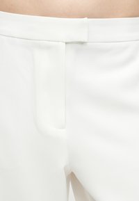 Gros plan sur un pantalon blanc ajusté avec une braguette avant dissimulée et une taille avec ceinture, porté sur une peau nue en arrière-plan.