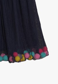 Laivansinisestä tyllihameesta, jonka helma on koristeltu värikkäillä pom-pomeilla pinkin, keltaisen ja sinisen sävyissä, lisäämään rakennetta ja visuaalista kiinnostavuutta alareunaan.