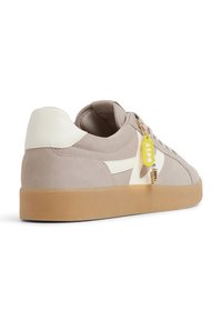 Sneaker in suede beige con dettagli in pelle bianca, suola in gomma, e un'etichetta gialla con un gancio, caratterizzato da un design low-profile e una superficie testurizzata.
