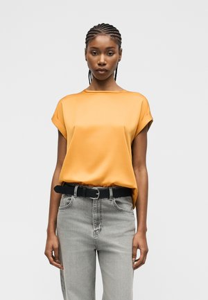 Kvinne med flettehår iført en sennepsgul kortermet bluse, lysegrå jeans og et svart belte, stående foran en ensfarget bakgrunn.