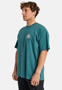 Teal cotton t-shirt with a crew neckline, short sleeves, and a graphic design featuring a triangle and text on the chest. Relaxed fit.