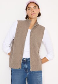 Beige geripptes ärmelloses Top mit hohem Kragen und durchgehendem Reißverschluss, getragen über einem weißen Langarmhemd und kombiniert mit blauen Jeans.