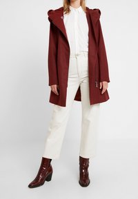 Burgundowa kurtka z kapturem z miękkiego materiału, z bocznymi zamkami, w połączeniu z kremowymi szerokimi spodniami i błyszczącymi brązowymi butami z tłoczonym wzorem krokodyla.