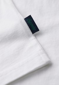Tela de algodón blanca con una textura suave. Presenta una etiqueta de un oscuro color navy que dice "Simply" en hilo verde. La camisa tiene costuras limpias y cosidas.