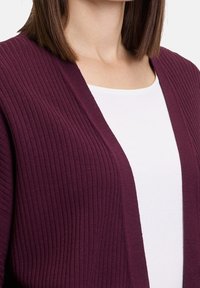 Žena oblečená v fialovém žebrovaném kardiganu přes jednoduché bílé triko s kulatým výstřihem, zobrazená od krku po střed trupu.