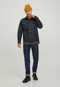 Veste en denim foncé avec un devant à boutons et des poches poitrine, associée à un jean foncé et des bottes marron. Les accessoires comprennent un bonnet en tricot jaune.