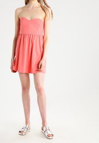 Korálové šaty bez ramínek s přiléhavým korzetem a rozšířenou sukní. Modelka má na sobě stříbrné skládací sandály. Hladká tkanina, minimalistický design.