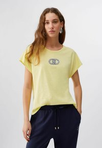 Jeune femme portant un t-shirt jaune clair avec un logo décoratif et un pantalon bleu marine à cordon, debout devant un fond blanc uni.