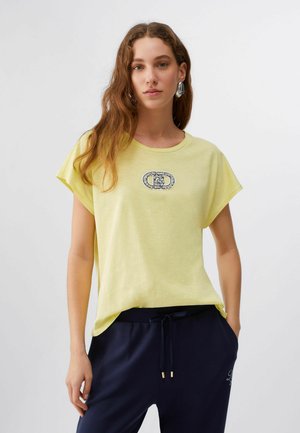 Giovane donna che indossa una maglietta giallo chiaro con logo decorativo e pantaloni con coulisse blu navy, in piedi davanti a uno sfondo bianco uniforme.