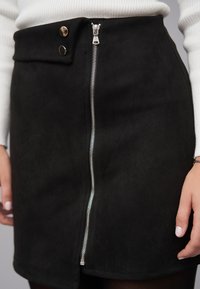Zwarte suède minirok met een rits aan de voorkant, twee drukknoopsluitingen ter hoogte van de taille en een gladde textuur. Getailleerde pasvorm met een rechte snit.