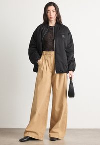 Sort bomberjakke med en logo patch, parret med beige brede bukser. Sorte spidse sko og en lille sort taske fuldender looket.