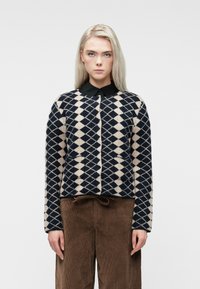 Mulher loira vestindo um suéter preto e bege com padrão de diamante sobre uma camisa preta com colarinho e calças de bombazine castanhas, de frente.