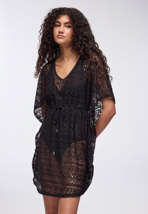 Czarna, lekka, szydełkowa sukienka narzutowa z dekoltem w serek, luźnymi rękawami, wiązaniem w talii oraz wzorem w zygzaki.