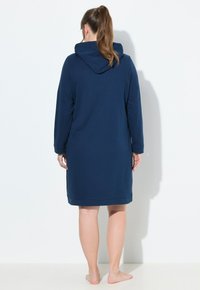 Donna in piedi a piedi nudi di spalle, indossando un abito blu navy con cappuccio, lungo fino al ginocchio e con maniche lunghe, contro uno sfondo bianco uniforme.