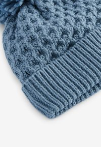 Bonnet en tricot bleu avec un motif texturé en point de câble et un bord côtelé. Présente un pompon doux et duveteux sur le dessus.