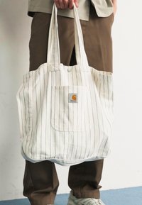 Bolsa de lona con rayas verticales, de color claro, diseño amplio, con un bolsillo frontal y parche con el logo, sostenida por una persona.