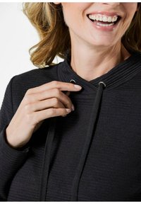 Zwarte pullover hoodie van een gestructureerde stof, met een funnel nek, koord met metalen oogjes en een close-up van een hand bij de halslijn.