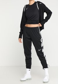 Czarny sportowy strój składający się z krótkiego topu, bluzy z kapturem na zamek oraz spodni z białymi paskami po bokach. Całość uzupełniają białe trampki.